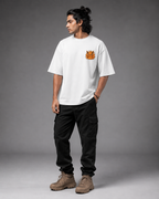 A man wearing a white crewneck t-shirt with a small orange graphic on the left chest, paired with black cargo pants and brown boots, standing against a gray studio background.