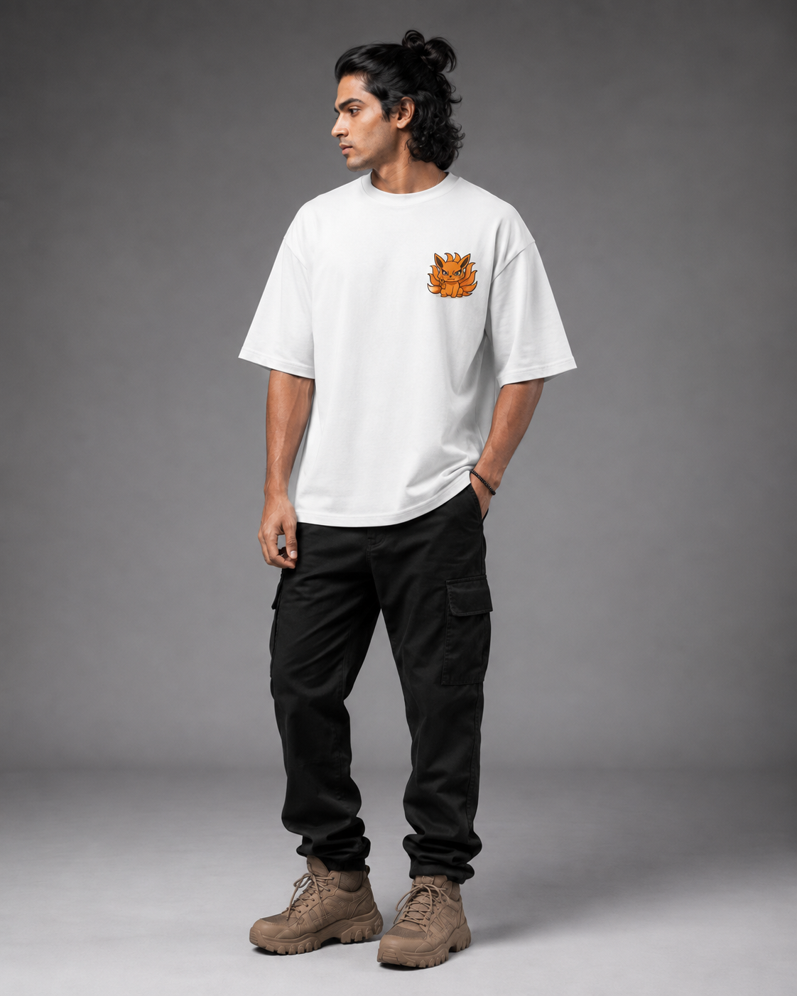 A man wearing a white crewneck t-shirt with a small orange graphic on the left chest, paired with black cargo pants and brown boots, standing against a gray studio background.