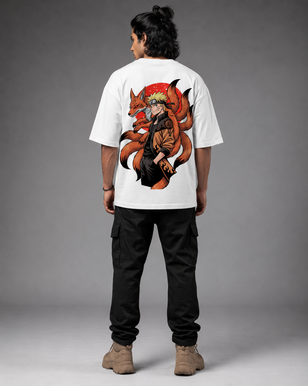 A man viewed from the back wearing a white oversized T-shirt with a large anime-style graphic on the back, paired with black cargo pants and tan boots, against a gray studio background.