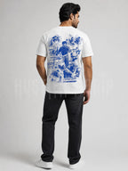 A man shown from the back wearing a white t-shirt with a large blue graphic print on the back, paired with black jeans and white sneakers, against a light gray studio background.