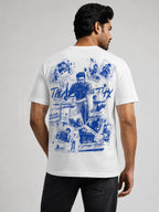A man shown from the back wearing a white t-shirt with a large blue graphic collage print on the back, paired with black jeans, against a light gray studio background.