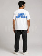 A man viewed from the back wearing a white t-shirt with a blue 'JANA MAYAGAN' graphic on the back, paired with black pants and white sneakers, against a light gray studio background.