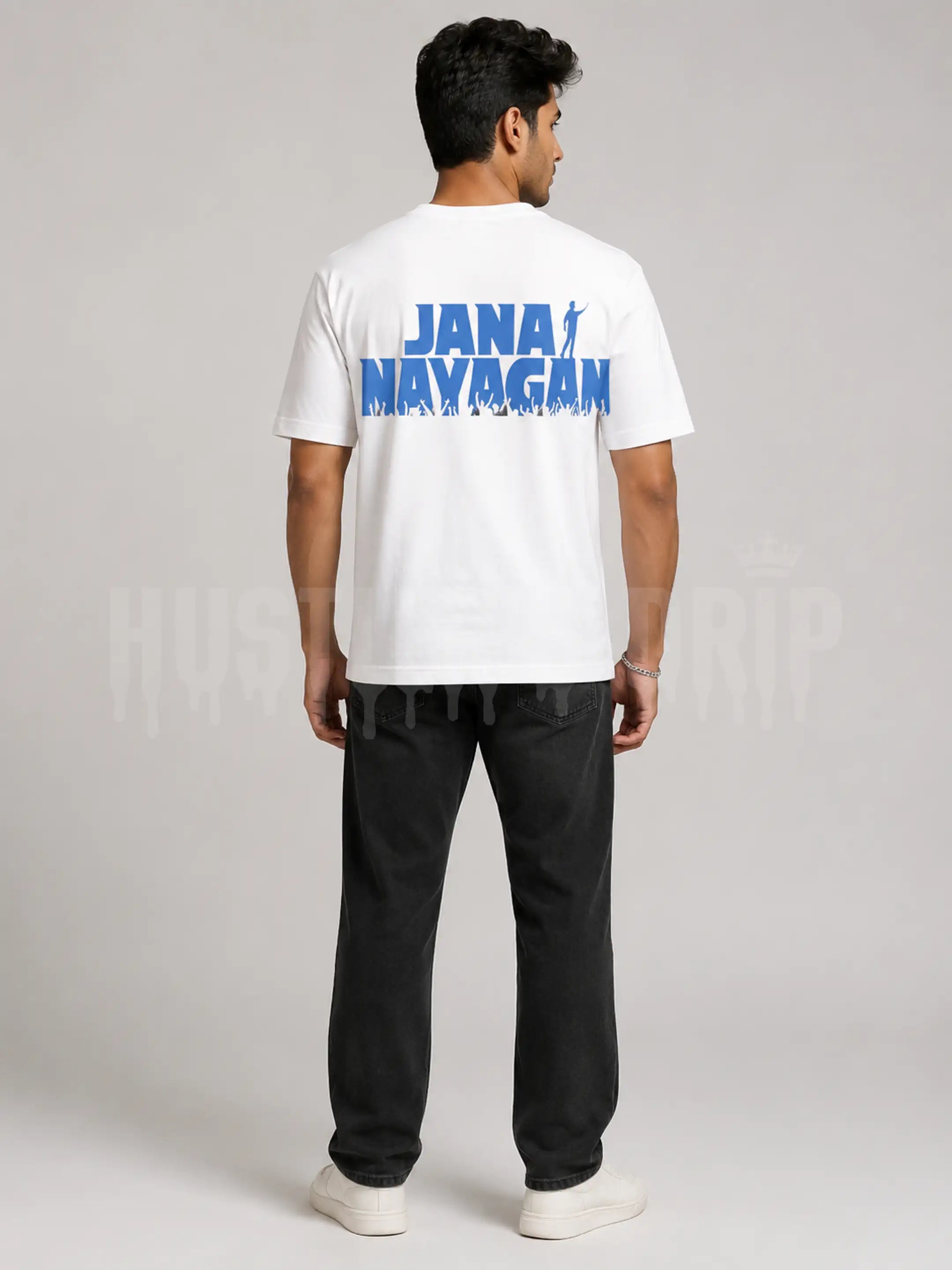 A man viewed from the back wearing a white t-shirt with a blue 'JANA MAYAGAN' graphic on the back, paired with black pants and white sneakers, against a light gray studio background.
