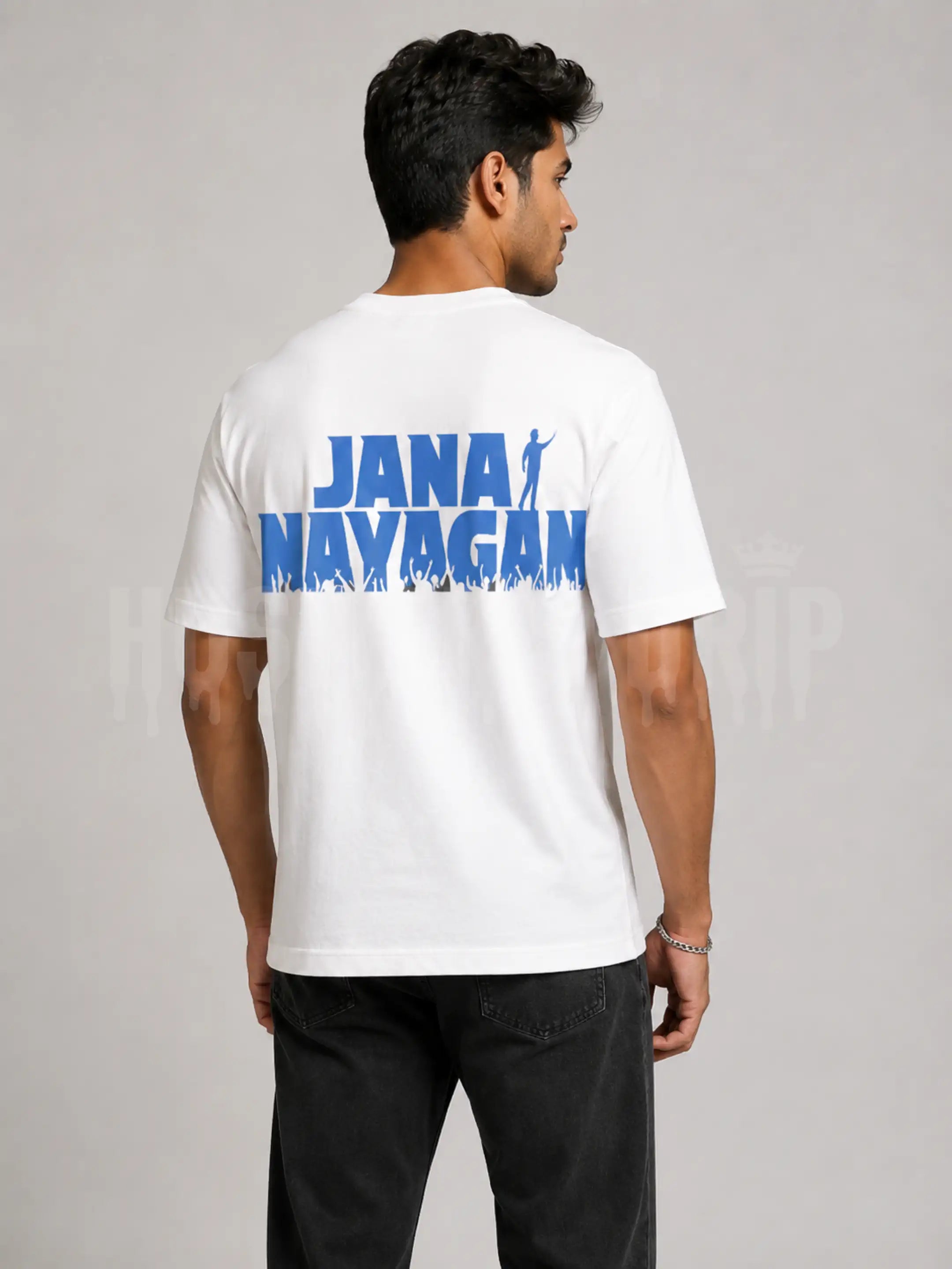 A man wearing a white t-shirt with a bold blue graphic print reading 'JANA NAYAGAN' on the back, shown from behind against a neutral gray studio background.