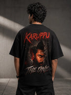 Person wearing a Suriya Karuppu black t-shirt with 'Karuppu The One' design on the back against a gray wall.
