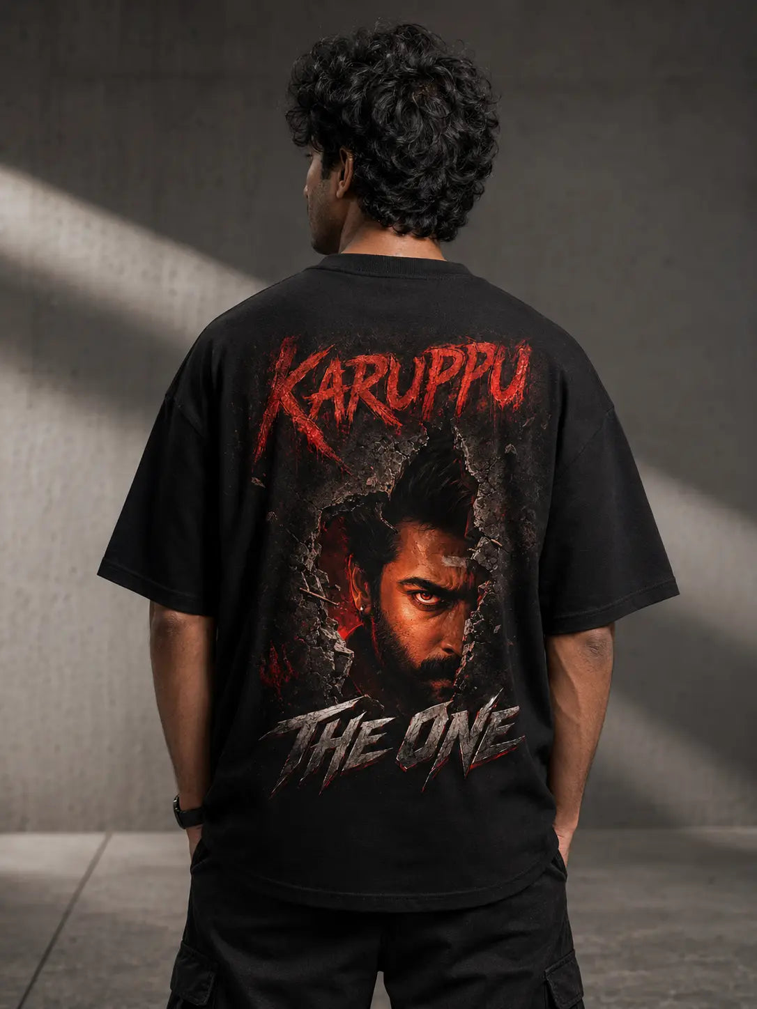 Person wearing a Suriya Karuppu black t-shirt with 'Karuppu The One' design on the back against a gray wall.
