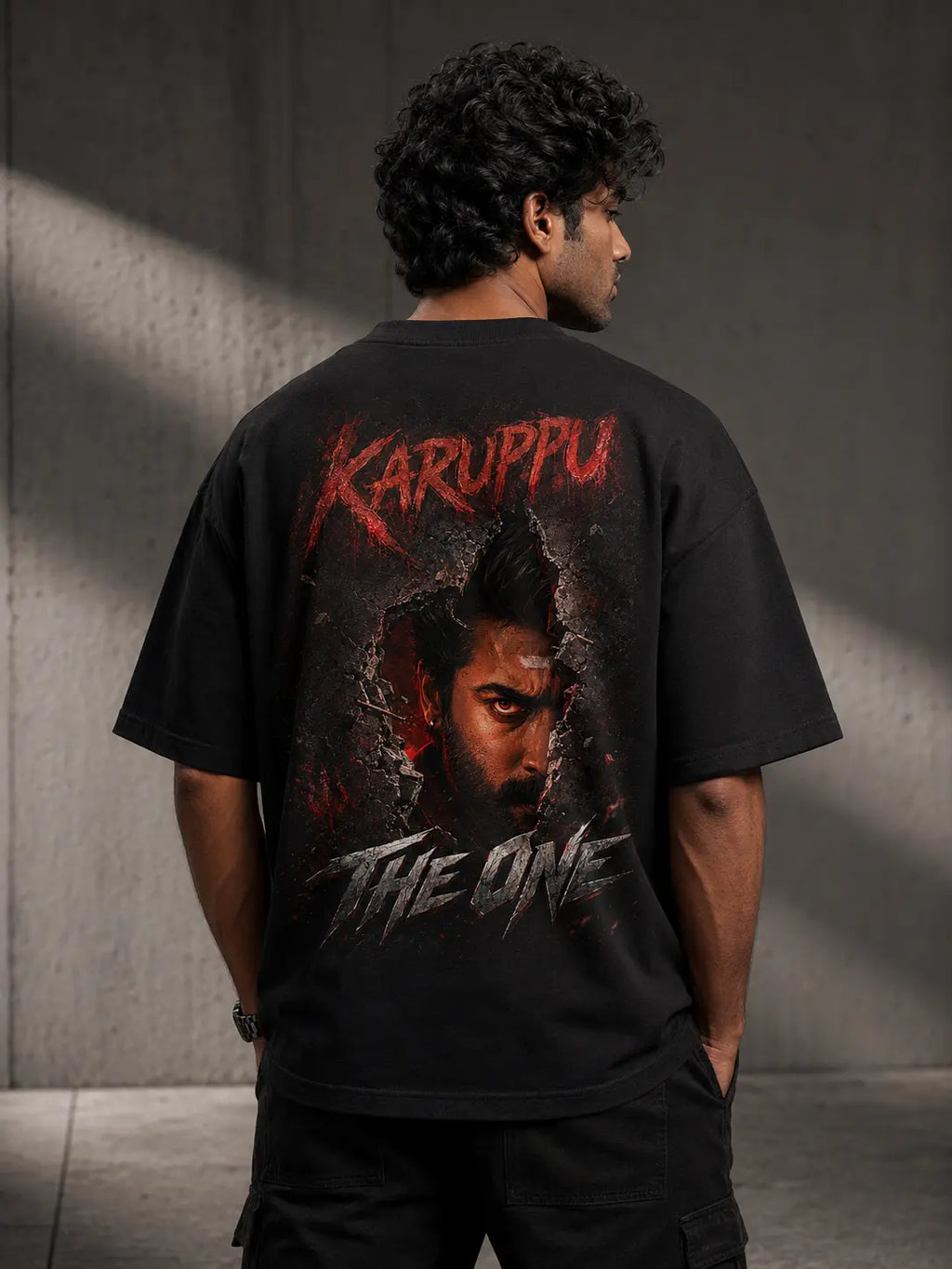 Person wearing a Suriya Karuppu black t-shirt with 'Karuppu The One' design on the back against a gray wall.