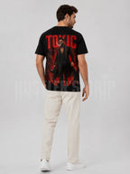A man wearing a black graphic t-shirt with a large red and black print on the back, paired with off-white trousers and white sneakers, shown from a rear three-quarter view against a light gray studio background.