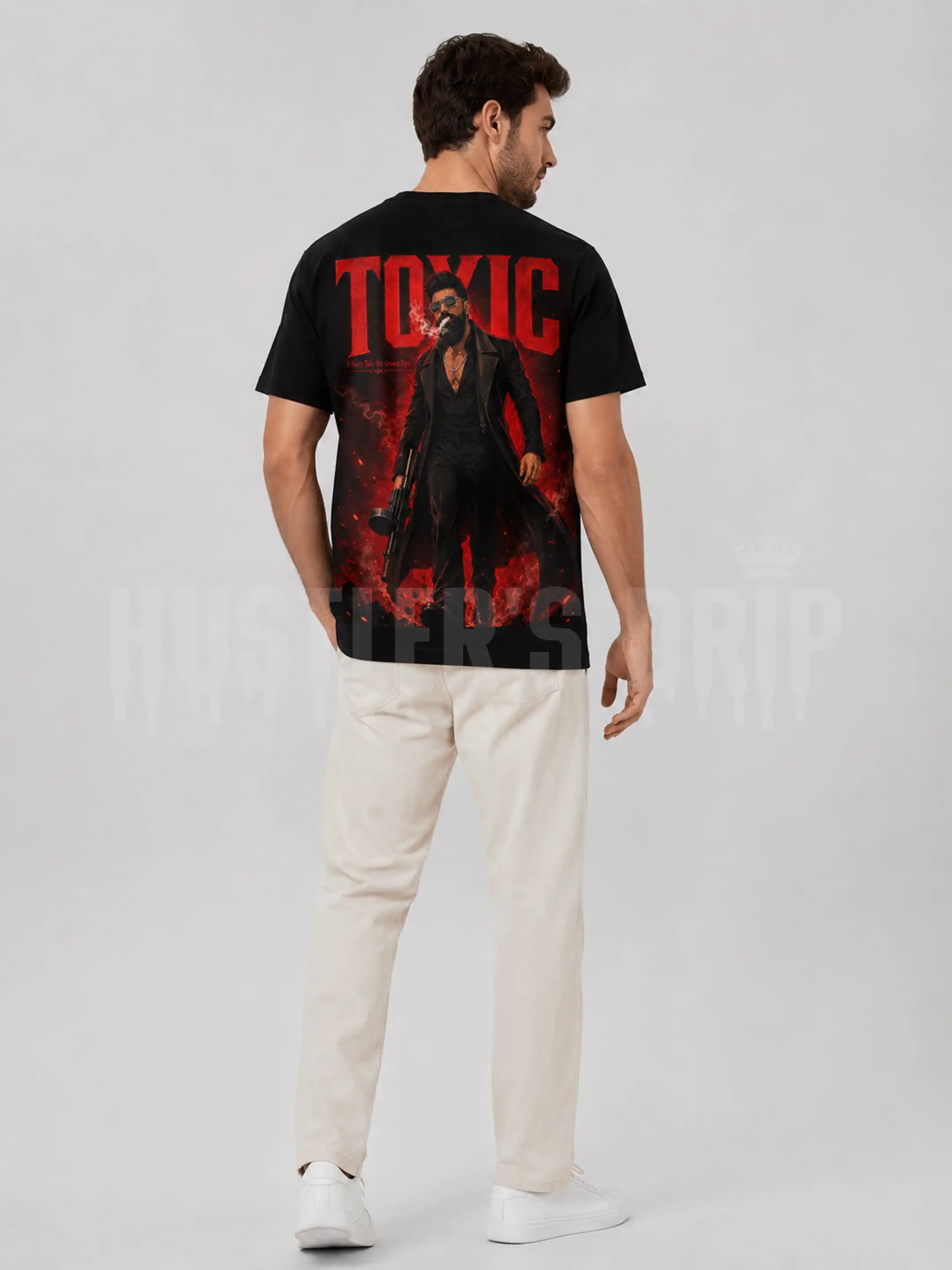A man wearing a black graphic t-shirt with a large red and black print on the back, paired with off-white trousers and white sneakers, shown from a rear three-quarter view against a light gray studio background.