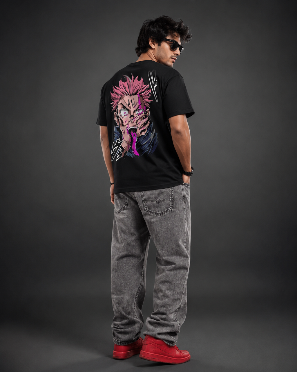 A man wearing a black graphic t-shirt with a large anime character print on the back, paired with gray jeans and red sneakers, photographed from behind against a dark gray studio background.