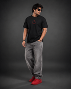 A man modeling a black crewneck t-shirt with a small red smiley face graphic on the chest, paired with gray jeans and red sneakers, set against a dark gray studio background.