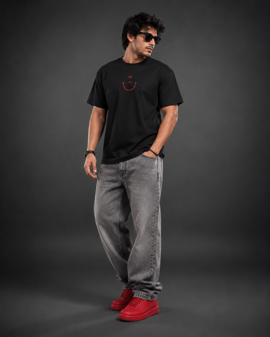 A man modeling a black crewneck t-shirt with a small red smiley face graphic on the chest, paired with gray jeans and red sneakers, set against a dark gray studio background.